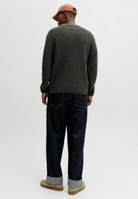 Man wearing rust-colored cap, dark green knit sweater, dark wide-leg jeans cuffed at ankles, and beige suede shoes, shown from behind.