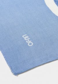 Foulard azzurro chiaro con una texture morbida, dotato di un logo bianco e un bordo leggermente sfrangiato, che mette in risalto un design semplice e moderno.