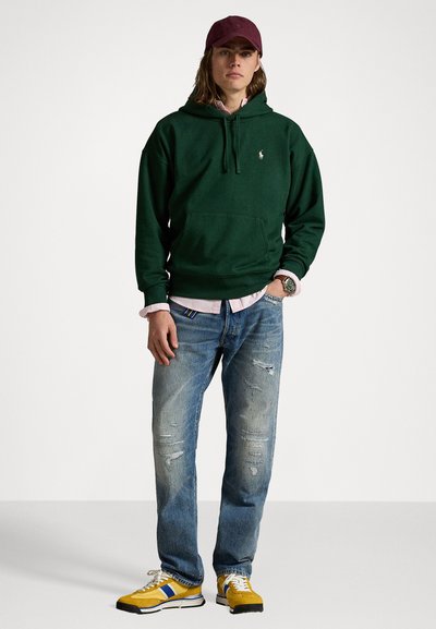 Grøn hættetrøje med en frontlomme, lagdelt over en lyserød ternet skjorte. Båret med slidte blå jeans og gule sneakers med blå detaljer.