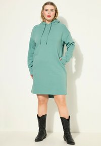 Teal šaty s kapucí, dlouhými rukávy, předn�ími kapsami a šňůrkou. Nosit s černými kovbojskými botami. Hladká textura látky.