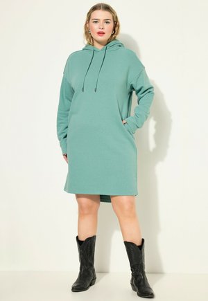 Vestido sudadera con capucha en color verde azulado, con mangas largas, bolsillos frontales y cordón ajustable. Usado con botas de cowboy negras. Textura de tela suave.