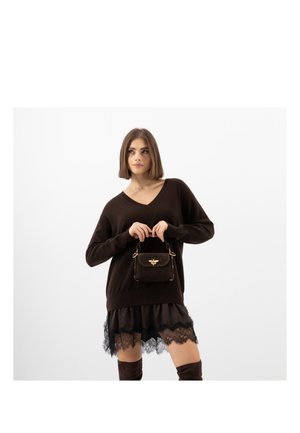 Vrouw die een donkerbruine trui-jurk met kanten rand draagt, een kleine donkerbruine handtas met een gouden bijenembleem vasthoudt, staand tegen een effen witte achtergrond.