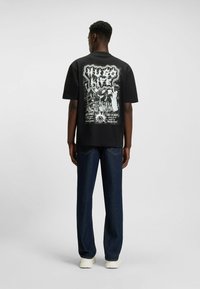 T-shirt noir avec imprimé graphique au dos, comportant des textes et illustrations blancs. Coupe ample, associé à un jean en denim bleu foncé et à des baskets blanches.