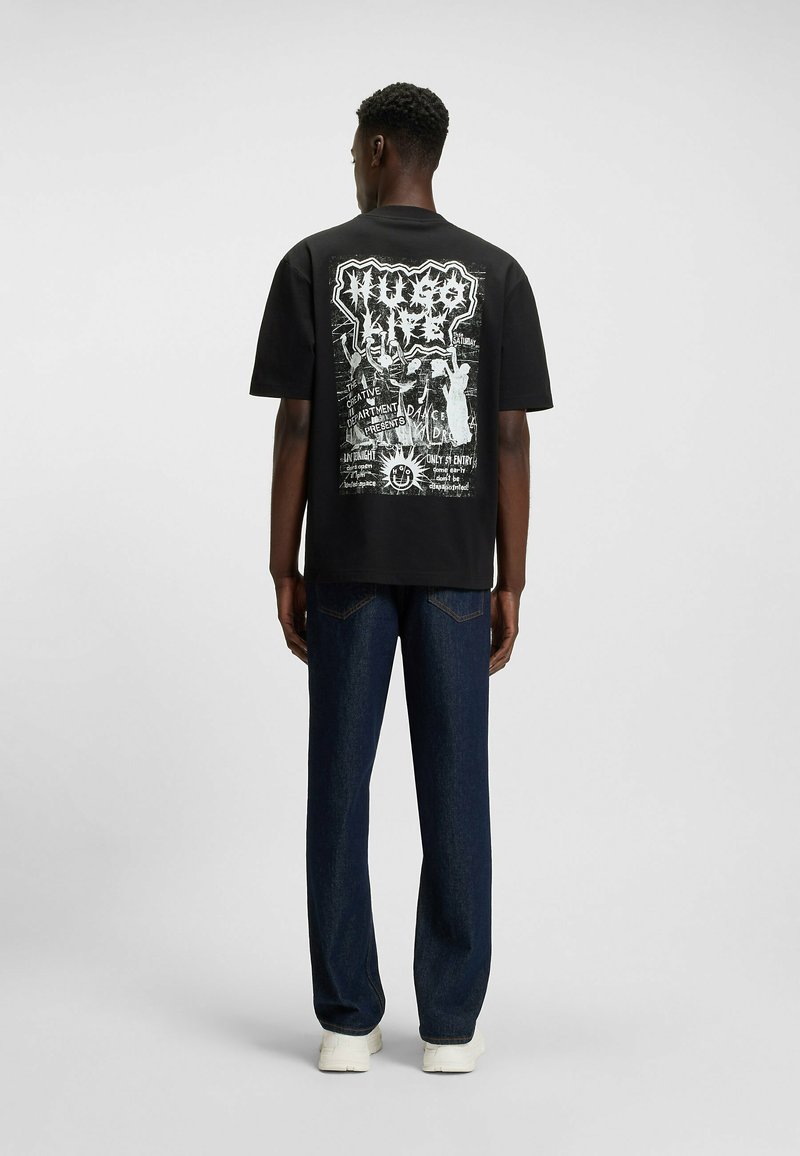 T-shirt noir avec imprimé graphique au dos, comportant des textes et illustrations blancs. Coupe ample, associé à un jean en denim bleu foncé et à des baskets blanches.