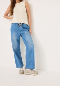 Haut sans manches beige clair en maille texturée, associé à un pantalon en denim bleu ample. Chaussures à lacets gris clair avec une semelle épaisse.