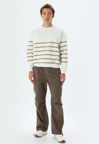 Sudadera blanca con rayas verde olivo, combinada con pantalones cargo marrón oscuro. El conjunto presenta un ajuste relajado y puños acanalados. Zapatillas blancas.