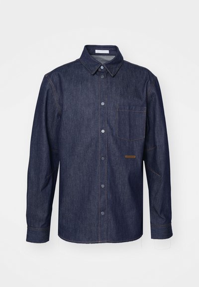 Sötét farmering, gombos elülső résszel, hegyes gallérral, egyetlen mellzsebbel és kontrasztos varrási részletekkel.