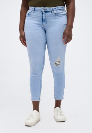 Jean skinny déchiré bleu clair porté avec des baskets blanches et un haut olive foncé, visible de la taille aux pieds sur un fond uni.