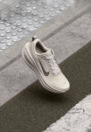 Beige Nike ZoomX hardloopschoen zweeft boven een gestructureerd trottoir en asfalt, met tastbare betegeling zichtbaar op de achtergrond.