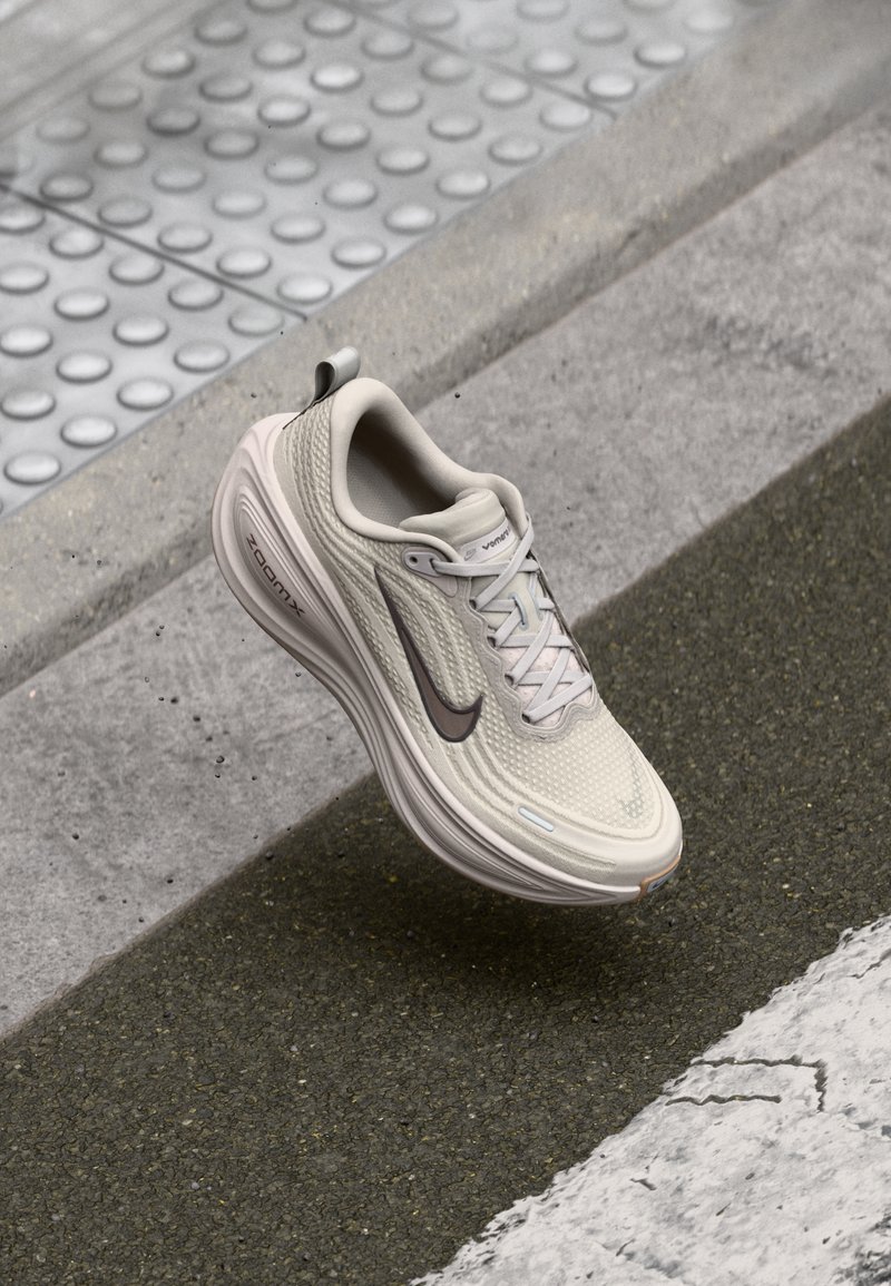 Beige Nike ZoomX running shoe suspended above textured sidewalk and asphalt with tactile paving visible in the background.