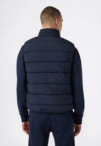 Mężczyzna w granatowej, pikowanej kamizelce oraz pasującym długim rękawie koszuli i spodniach, stojący tyłem na jednolitym tle.