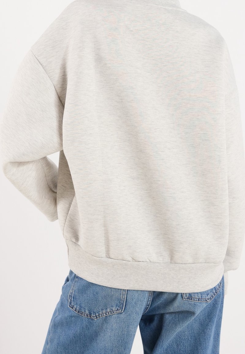 Personne portant un sweat-shirt gris clair et un jean en denim bleu, vue de dos avec une main posée sur la hanche.