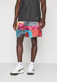 Mand iført multikolorede basketball-print shorts, hvide Puma sneakers, hvide sokker og en sort mesh ærmeløs top, stående foran en ensfarvet baggrund.