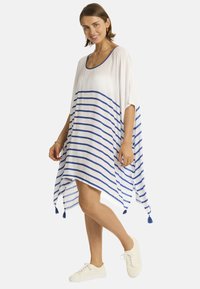 Kaftano bianco con righe orizzontali blu, caratterizzato da una scollatura rotonda, maniche svasate e dettagli con nappine all'orlo. Indossato con sneakers bianche.