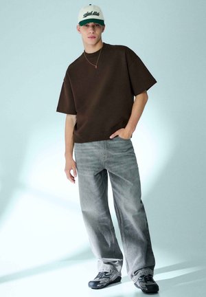 Junger Mann trägt ein dunkelbraunes, übergroßes T-Shirt, weite graue Jeans, schwarze und graue Sneakers sowie eine weiße Baseballkappe mit grüner Schrift.