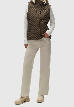 Osoba nosící hnědou prošívanou vestu s potiskem přes bílé tričko s dlouhým rukávem, béžové kalhoty s širokými nohavicemi a béžové robustní boty.