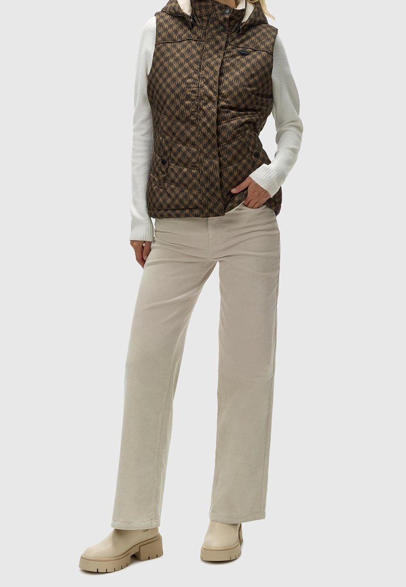 Personne portant un gilet matelassé marron à motif par-dessus un top blanc à manches longues, un pantalon beige à jambes larges et des bottes beiges épaisses.