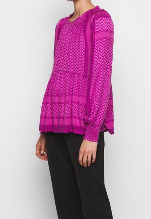 Femme portant un chemisier à manches longues rose vif à motifs avec un pantalon noir, montrée des épaules aux hanches sur un fond uni clair.