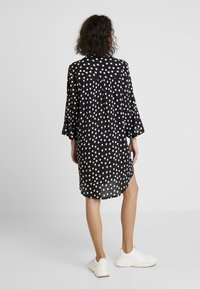 Schwarzes Polka-Dot-Kleid mit Dreiviertelärmeln, lockerer Passform und asymmetrischem Saum, aus leichtem Stoff. Getragen mit weißen Sneakern.