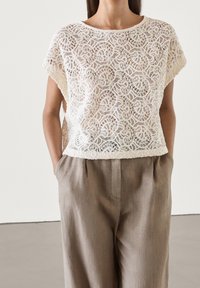 Haut en dentelle écru avec un col rond et des manches courtes, présentant un motif floral complexe. Associé à un pantalon en lin beige.