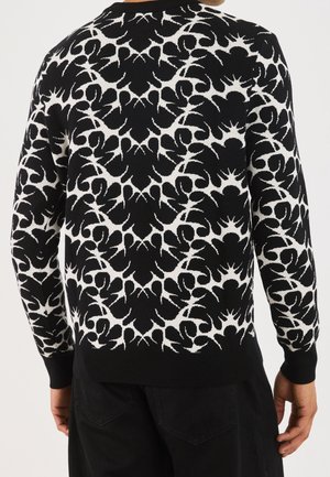 Sort-hvid langærmet sweater med symmetrisk abstrakt stjerneskud- og blomstermønster på en person, der står med ryggen mod kameraet.