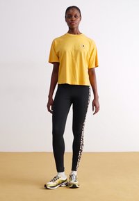 Sárga rövidített póló, fekete leggings oldalán fehér mintával, és sárga sportcipő. A modell egy egyszerű háttér előtt áll.