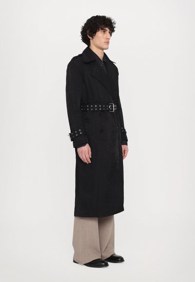 Schwarzer Trenchcoat mit Gürtel und Metallösen, doppelreihiger Schnitt und hochgeschlagenen Ärmeln. Trägt man über beigen weiten Hosen.