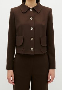 Brun kort jacka med rund krage, försedd med sex metallknappar och två framfickor. Slät tyg med strukturerad design.