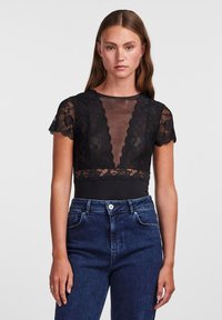 Body nero con maniche corte in pizzo, pannello frontale trasparente, bordi ondulati, abbinato a jeans blu a vita alta.