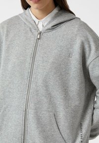 Szara bluza z kapturem zapinana na zamek, wykonana z miękkiego materiału. Posiada przednią kieszeń oraz haftowane logo na piersi.