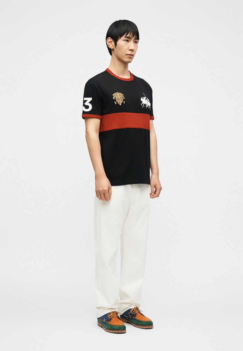 Homme portant un polo à rayures noires et rouges avec le numéro blanc 3 sur la manche, un pantalon blanc, et des chaussures multicolores bleu, orange et vert.