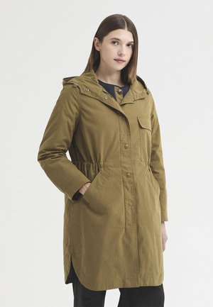 Femme aux cheveux bruns lisses portant un long manteau à capuche vert olive avec des poches avant et des fermetures par boutons, debout devant un fond uni.
