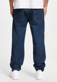 Dark blue denim jeans with a relaxed fit, featuring back pockets and orange stitching. Paired with white athletic shoes.