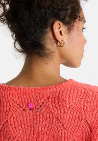 Korallenfarbener gestrickter Pullover mit strukturiertem Muster, Rundhalsausschnitt und pinkem Knopfdetail im Nacken. Weiches, flauschiges Material.