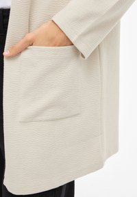 Ανοιχτό μπεζ πλεκτή ζακέτα με ορατή τσέπη, που διαθέτει διακριτικό κυματιστό σχέδιο και άνετη εφαρμογή. Μαλακό υλικό, μανίκια τριών τετάρτων.