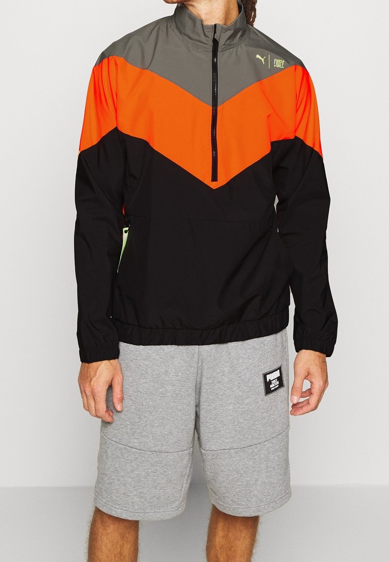 Man som bär en färgblockerad jacka i svart, orange och grått med dragkedja samt grå Puma-shorts, stående mot en enkel ljus bakgrund.