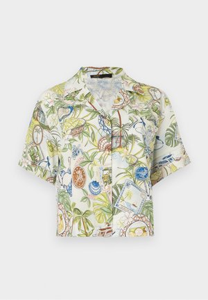 Kobieta stoi zwrócona przodem, ubrana w krótki, kwiecisty koszulowy t-shirt z guzikami, beżowe szorty z wysokim stanem, białe skarpetki i brązowe buty na platformie.