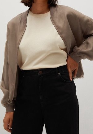Personne portant un pull léger beige, une veste bomber taupe avec des manches transparentes et un pantalon noir taille haute, main posée sur la hanche.