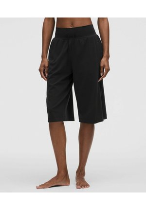 Personne portant un short noir ample jusqu'aux genoux avec une taille à cordon, debout pieds nus devant un fond clair uni.