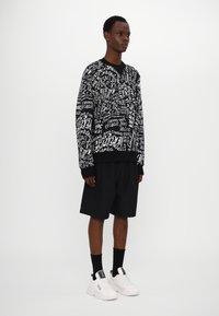 Dugi sweatshirt u crnoj i bijeloj boji, s raznim uzorcima teksta; uparen s crnim šortsevima i bijelim tenisicama.
