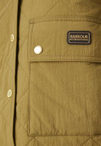 Kurtka pikowana w kolorze oliwkowym, z naszywaną kieszenią, złotymi zatrzaskami oraz czarną metką z białym napisem "BARBOUR INTERNATIONAL."