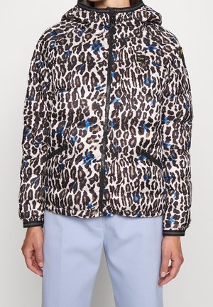 Kurtka typu puffer w panterkę z niebieskimi akcentami, czarnym zamkiem i kieszeniami, noszona z jasnoniebieskimi spodniami.