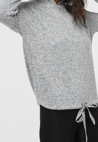 Femme portant un sweat à capuche gris clair à motifs, avec une poche avant et un cordon de serrage réglable, associé à des leggings noirs.