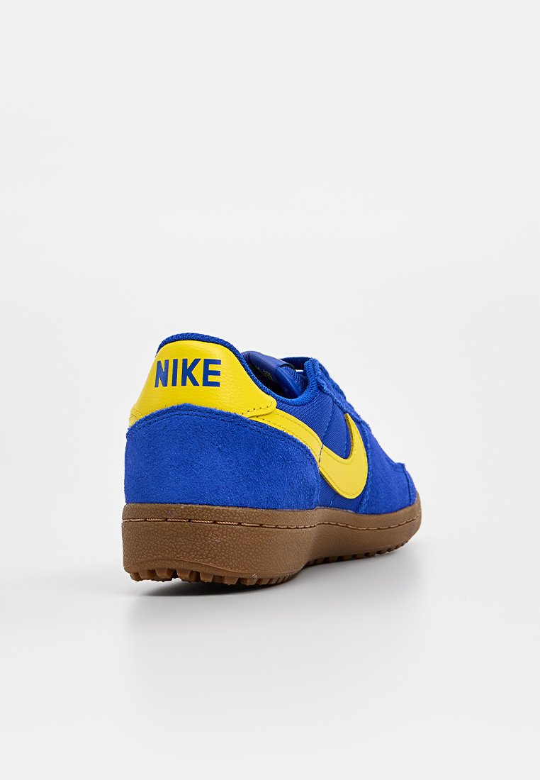 Blaue Wildleder-Sneaker mit gelben Akzenten, die ein markantes gelbes Nike-Logo an der Ferse und eine braune Gummisohle aufweisen. Abgerundete Form.