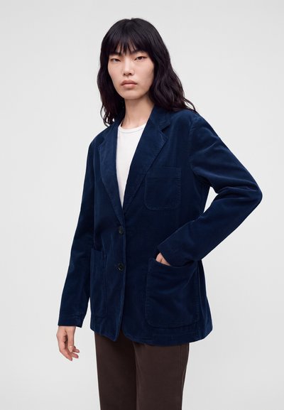 Personne aux longs cheveux noirs portant un blazer en velours côtelé bleu marine sur une chemise blanche et un pantalon sombre, debout devant un fond clair uni.
