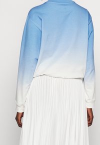 Sudadera azul clara con un efecto degradado que se desvanece hasta el blanco. Presenta mangas largas y un escote redondeado, combinada con una falda blanca plisada.