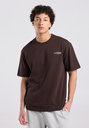Jonge man met krullend haar, gekleed in een donkerbruin Lazy&Broke T-shirt en lichtgrijze broek, staand tegen een eenvoudige lichte achtergrond.