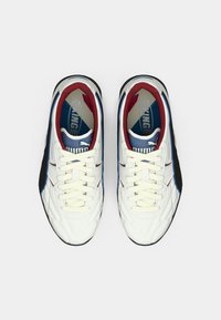 Um par de ténis brancos da Puma com atacadores creme, forro interior vermelho, detalhes azuis e pretos, vistos de cima sobre um fundo branco.