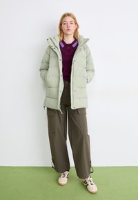 Giacca puffer verde chiaro con cappuccio, bottoni e tasche laterali; abbinata a un top viola scuro e pantaloni cargo oliva. Presenta sneakers.