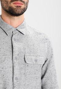 Šedá košile na knoflíky vyrobená z texturované tkaniny, s tečkovaným vzorem, límcem a dvěma náprsními kapsami se zapínáním na knoflíky.